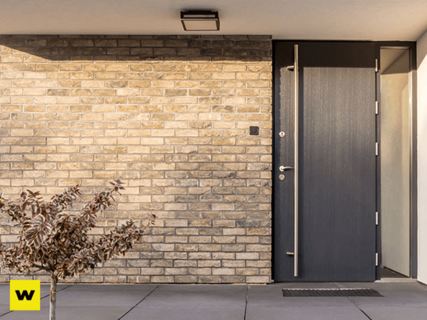 Moderne Haustür aus Aluminium vor heller Klinkerfassade mit kleinem Baum und Licht.